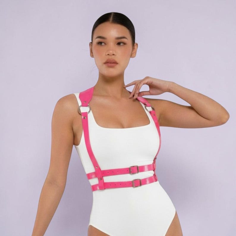 Pu&ograve; includere: Un'imbracatura rosa acceso con anelli e fibbie in metallo color argento. Indossata sopra un body bianco. Le cinghie passano sulle spalle e intorno alla vita. Lo sfondo &egrave; di colore viola chiaro.