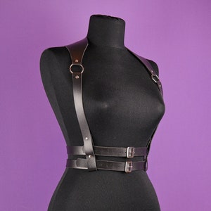 Pu&ograve; includere: Imbracatura per il corpo in pelle nera con elementi metallici color argento. L'imbracatura presenta spallacci collegati da anelli metallici, una cintura con due cinturini regolabili e fibbie color argento. Esposta su un manichino nero su sfondo viola.