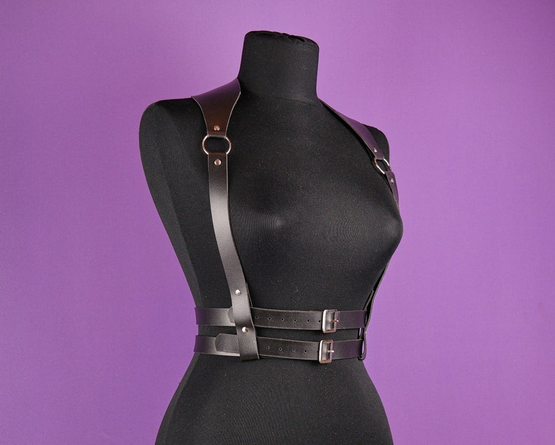 Pu&ograve; includere: Imbracatura per il corpo in pelle nera con elementi metallici color argento. L'imbracatura presenta spallacci collegati da anelli metallici, una cintura con due cinturini regolabili e fibbie color argento. Esposta su un manichino nero su sfondo viola.