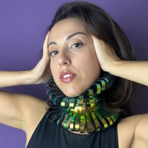 May include: A statement necklace with a layered design. The necklace features metallic gold and green elements, creating a striking visual effect. The piece is worn against a black top, with a purple background.