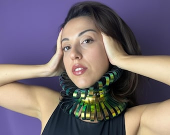 Op de afbeelding: Een opvallende statement ketting met een gelaagd ontwerp. De ketting heeft metallic gouden en groene elementen, wat een opvallend visueel effect creëert. Het sieraad wordt gedragen op een zwarte top, met een paarse achtergrond.