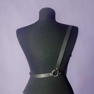 May include: Black leather body harness with a single shoulder strap and a waist belt. The harness features a silver-colored metal ring and is displayed on a black mannequin against a purple background.