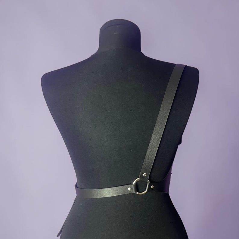 May include: Black leather body harness with a single shoulder strap and a waist belt. The harness features a silver-colored metal ring and is displayed on a black mannequin against a purple background.