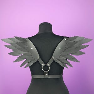 May include: Black faux leather shoulder harness with layered feather-like wings. The harness features a central metal ring and adjustable straps. Silver-toned rivets secure the wing attachments. The wings are a dark gray color, and the background is a solid purple.