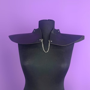 Puede incluir: Un collar de cuero morado oscuro con una cadena plateada, exhibido en un maniquí negro. El collar tiene un diseño ancho y angular con remaches en los bordes. El fondo es de color morado liso.