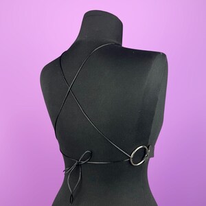 K&ouml;nnte beinhalten: Schwarzes Neckholder-Top mit gekreuztem R&uuml;ckendesign. Das Top hat ein silberfarbenes Ringdetail und einen Kordelverschluss am unteren R&uuml;cken. Das Kleidungsst&uuml;ck wird auf einer schwarzen Schaufensterpuppe vor lila Hintergrund pr&auml;sentiert.