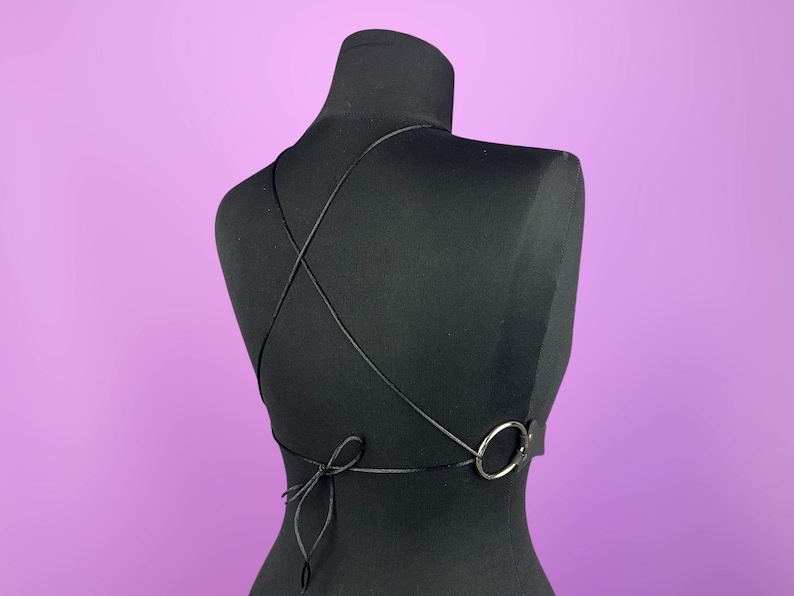 K&ouml;nnte beinhalten: Schwarzes Neckholder-Top mit gekreuztem R&uuml;ckendesign. Das Top hat ein silberfarbenes Ringdetail und einen Kordelverschluss am unteren R&uuml;cken. Das Kleidungsst&uuml;ck wird auf einer schwarzen Schaufensterpuppe vor lila Hintergrund pr&auml;sentiert.
