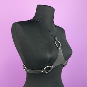 Puede incluir: Arn&eacute;s de cuerpo de cuero negro con anillos y herrajes de metal plateado. El arn&eacute;s presenta un dise&ntilde;o de cuello halter y correas ajustables. El producto se exhibe en un maniqu&iacute; negro sobre un fondo p&uacute;rpura.