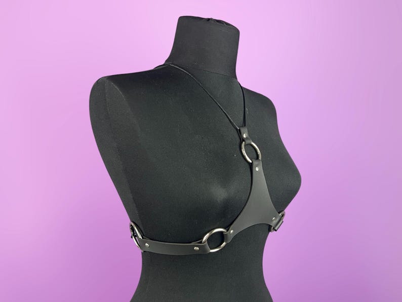 Puede incluir: Arn&eacute;s de cuerpo de cuero negro con anillos y herrajes de metal plateado. El arn&eacute;s presenta un dise&ntilde;o de cuello halter y correas ajustables. El producto se exhibe en un maniqu&iacute; negro sobre un fondo p&uacute;rpura.