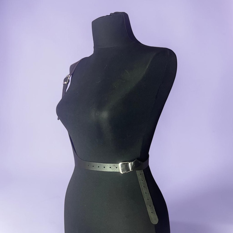 May include: A black leather body harness displayed on a black mannequin. The harness features a thin strap that goes over one shoulder and a belt that wraps around the waist. The belt has a silver buckle. The background is a solid light purple.