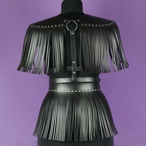May include: Black leather harness with fringe detail. The harness has a wide belt with a metal ring and studs. The fringe is attached to the belt and the shoulders.