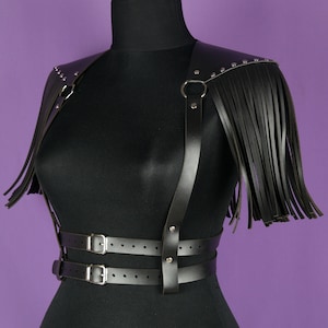 May include: Black leather harness with fringe detail and silver hardware. The harness has a wide belt with two buckles and adjustable straps that go over the shoulders.