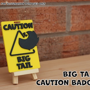 May include: Yellow caution sign with black text and a black silhouette of a tail. The sign reads "CAUTION BIG TAIL" and is displayed on a small wooden easel. The sign is a warning badge.