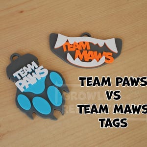 May include: Two gray plastic dog tags with the text "TEAM PAWS" and "TEAM MAWS" in white and orange letters. The "TEAM PAWS" tag is shaped like a paw print with a blue paw pad. The "TEAM MAWS" tag is shaped like a set of teeth.