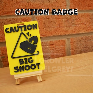 May include: Yellow and black caution sign with a black triangle and the text "CAUTION BIG SNOOT" on a small wooden easel.