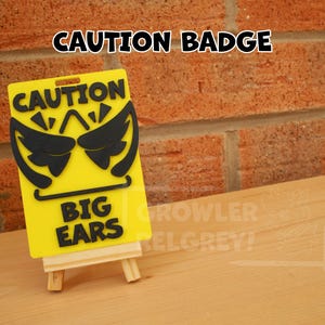 May include: Yellow and black caution sign with the text "CAUTION BIG EARS" and a cartoon image of large ears. The sign is on a small wooden easel.