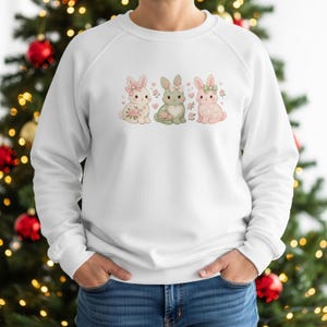 May include: White sweatshirt featuring three cartoon bunnies in pastel colors. The bunnies have floral accents and bows. The sweatshirt is a crew neck style and is made of a soft material. The design is centered on the front of the sweatshirt.