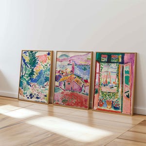Könnte beinhalten: Drei gerahmte abstrakte Landschaftsbilder. Das Kunstwerk zeigt leuchtende Farben, darunter Blau, Grün, Rosa und Gelb. Die Gemälde zeigen Szenen von Natur, Gebäuden und Küstenansichten. Die Rahmen sind hellbraun und lehnen an einer weißen Wand.