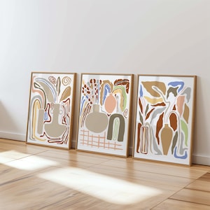 Könnte beinhalten: Drei abstrakte Kunstdrucke im modernen minimalistischen Stil. Jeder Druck zeigt eine andere Anordnung von organischen Formen und Linien in Braun-, Beige-, Pfirsich-, Blau- und Grüntönen. Die Drucke sind in Naturholz gerahmt.