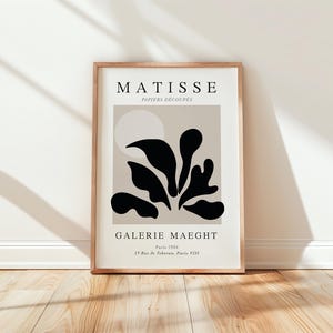 Puede incluir: Una impresión enmarcada de una obra de arte de Matisse titulada "Papiers Decoupes" con un diseño abstracto negro sobre un fondo beige. La impresión incluye el texto "MATISSE", "GALERIE MAEGHT", "París 1954" y "13 Rue de Téhéran, París VIII".
