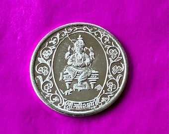 May include: A silver coin featuring an image of a seated deity with multiple arms, wearing a crown, and surrounded by an ornate border. The coin is set against a vibrant magenta background.