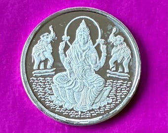 May include: Silver coin featuring a seated deity with a halo, flanked by elephants. The coin has intricate details and is set against a purple background. The design is embossed with a detailed pattern.