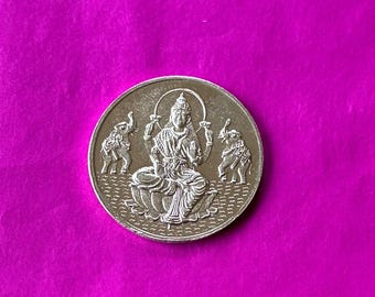 May include: A silver coin featuring a seated deity with a halo, flanked by two elephants. The coin is set against a vibrant magenta background, showcasing intricate details of the religious figure and the elephants.