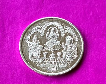 May include: A silver coin with intricate carvings of deities, set against a vibrant magenta background. The coin features detailed figures within an arched design, with text at the bottom. The coin's design suggests a religious or cultural theme.