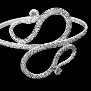 May include: A silver-toned arm cuff with an open design. The cuff features a textured, snake-like pattern that wraps around the arm. The design includes two large, looping swirls, creating a unique and elegant accessory.