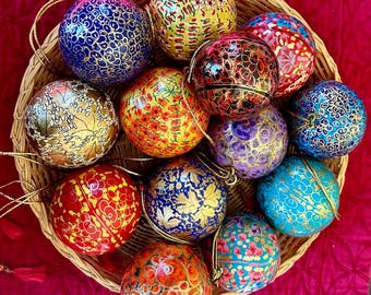 Puede incluir: Una colección de 12 adornos navideños de papel maché pintados a mano en una cesta de mimbre. Los adornos están decorados con intrincados patrones en oro, rojo, azul, verde y morado.