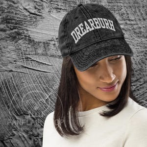 May include: A black baseball cap with the word "DREARBURH" in white block letters. The cap has a curved brim and a distressed, vintage look. The cap is worn by a person.