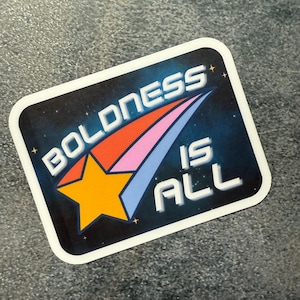 May include: A rectangular sticker with rounded corners. The sticker features the text "BOLDNESS IS ALL" in white, set against a dark blue background with stars. A yellow star and colorful comet streaks are also present.