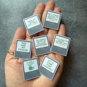 May include: A collection of gray rectangular stickers with white borders and green text. The stickers feature various phrases like "You are my friend" and "It's time for lasers!" The word "RATCHED" is printed on each sticker.