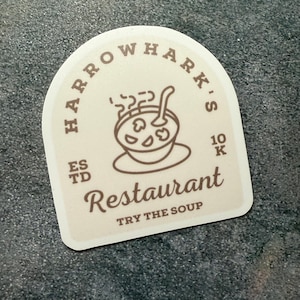 May include: Cream-colored sticker with rounded corners featuring the text "HARROWHARK'S" in a curved font. Below, the word "Restaurant" is written in a cursive font, with "TRY THE SOUP" underneath. A soup bowl graphic is in the center.