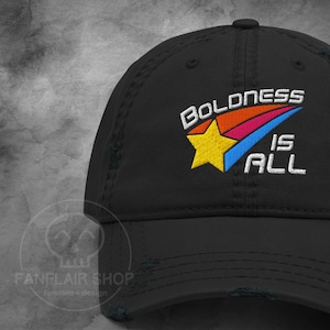 以下が含まれることがあります： 黒色の野球帽。刺繍された「BOLDNESS IS ALL」の文字が白で、黄色の星とカラフルな矢印のデザインが特徴です。Fanflair Shopの商品です。