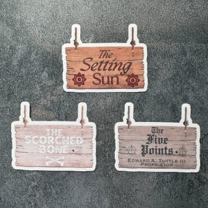 May include: Three sticker signs with a rustic wood design and rope hangers. The signs read "The Setting Sun", "The Scorched Bone", and "The Five Points". The Five Points sign includes "Edward A. Tuttle III Proprietor".