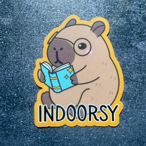 May include: A sticker featuring a cartoon capybara wearing glasses and reading a book with blue pages and gold stars. The sticker has a yellow border and the word "INDOORSY" in white block letters.