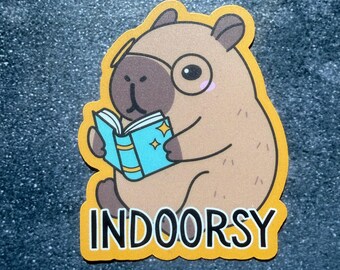 May include: A sticker featuring a cartoon capybara wearing glasses and reading a book with blue pages and gold stars. The sticker has a yellow border and the word "INDOORSY" in white block letters.
