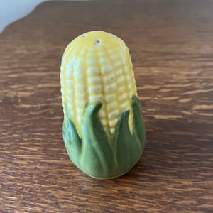 May include: A ceramic corn on the cob salt shaker with green leaves. The salt shaker is yellow and has a hole on the top for pouring.