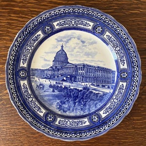 May include: A blue and white decorative plate with a detailed illustration of the U.S. Capitol building. The plate has a scalloped edge and ornate border designs. The central image depicts the Capitol with trees and a cloudy sky.