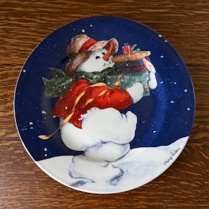 May include: A decorative plate featuring a snowman carrying gifts. The snowman wears a brown hat, red coat, and green scarf against a dark blue background with falling snow. The plate has a white rim.