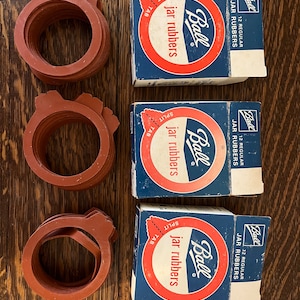 May include: Three boxes of Ball brand jar rubbers, each containing 12 regular jar rubbers. The boxes are blue and red with white text. The text on the boxes reads "Ball jar rubbers" and "12 regular jar rubbers". There are three stacks of red rubber jar rings next to the boxes.