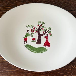 May include: White oval ceramic plate with a hand-painted design of a green tree with red apples, a small house, and two people in red and green clothing.
