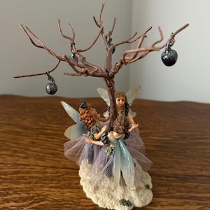 May include: A whimsical fairy figurine featuring three fairies beneath a copper-colored tree with hanging dark purple orbs. The fairies have delicate wings and are dressed in pastel-colored gowns. The base is a textured, off-white color.