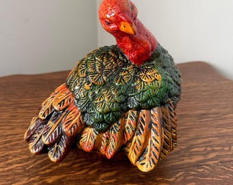May include: A ceramic turkey figurine featuring a bright red head and neck with a yellow beak. The body is covered in textured, overlapping feathers painted in shades of deep forest green, burnt orange, and brown with a glossy finish.