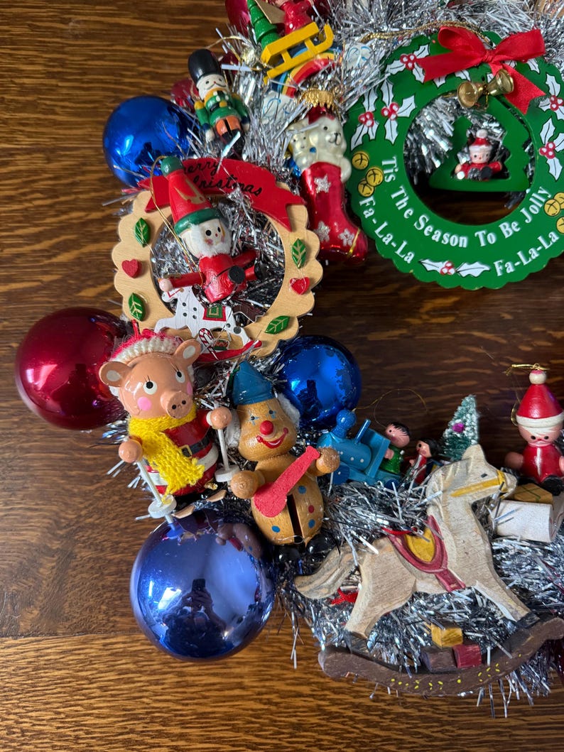 May include: A festive Christmas wreath with various ornaments, including a pig in a yellow scarf, a clown, a rocking horse, and a nutcracker. The wreath includes a green circular sign that reads "Tis The Season To Be Jolly".