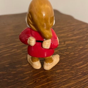 Może przedstawiać: Ceramiczna figurka krasnala w czerwonej kurtce i brązowym kapeluszu. Krasnal stoi z założonymi rękami i jest odwr&oacute;cony plecami do kamery.