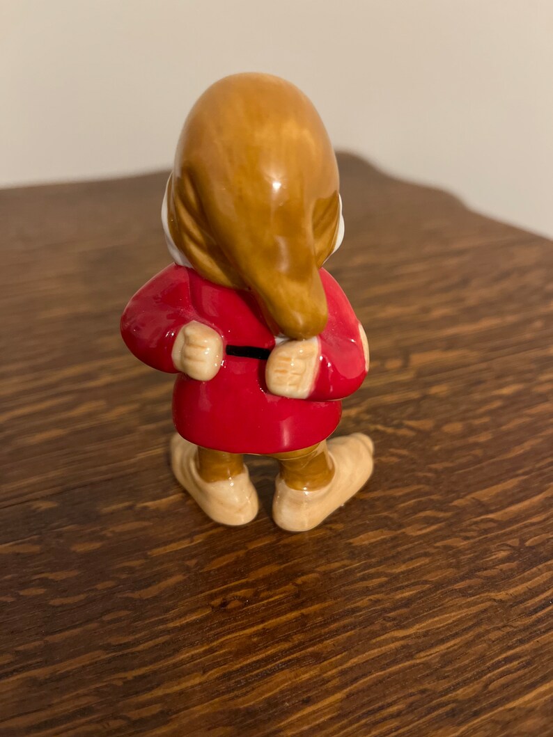 Może przedstawiać: Ceramiczna figurka krasnala w czerwonej kurtce i brązowym kapeluszu. Krasnal stoi z założonymi rękami i jest odwr&oacute;cony plecami do kamery.