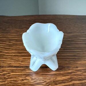 May include: A small, opalescent white glass bowl with a decorative, floral-like design. The bowl is supported by three curved legs, creating a pedestal effect. The bowl's rim has a scalloped edge.