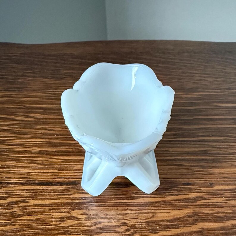May include: A small, opalescent white glass bowl with a decorative, floral-like design. The bowl is supported by three curved legs, creating a pedestal effect. The bowl's rim has a scalloped edge.